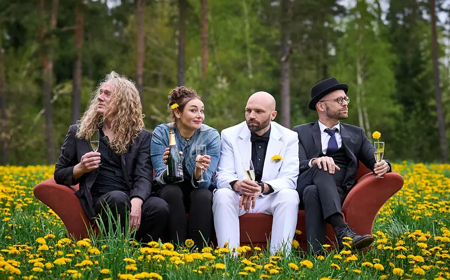 Vier Personen sitzen auf einem Sofa in einer Blumenwiese und halten Gläser; im Hintergrund ein Wald.