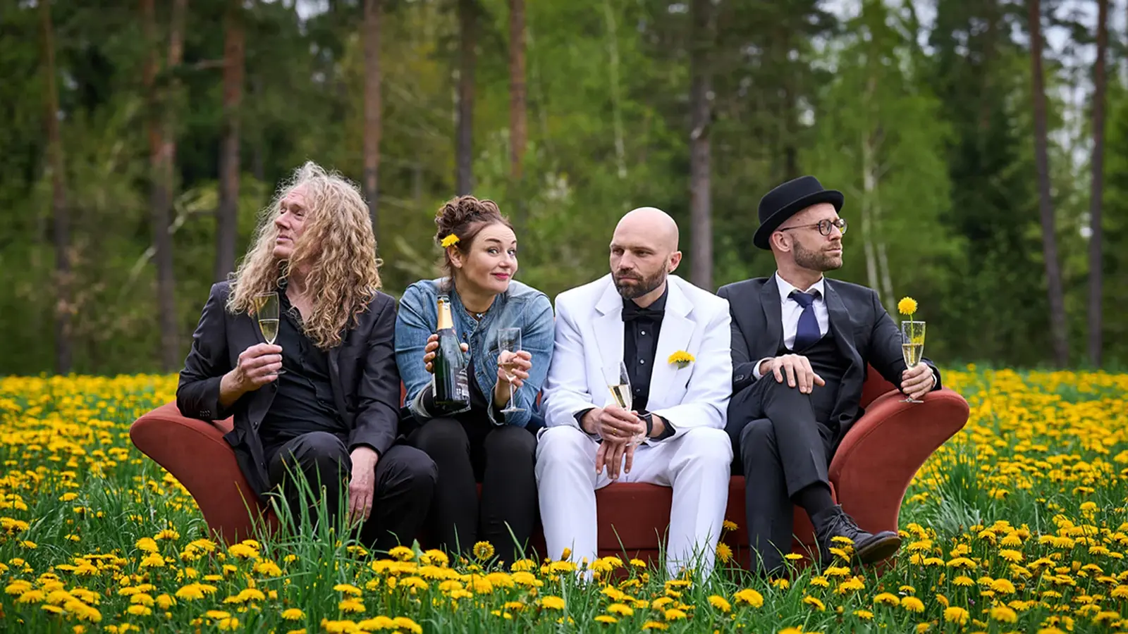 Vier Personen sitzen auf einem Sofa in einer Blumenwiese und halten Gläser; im Hintergrund ein Wald.