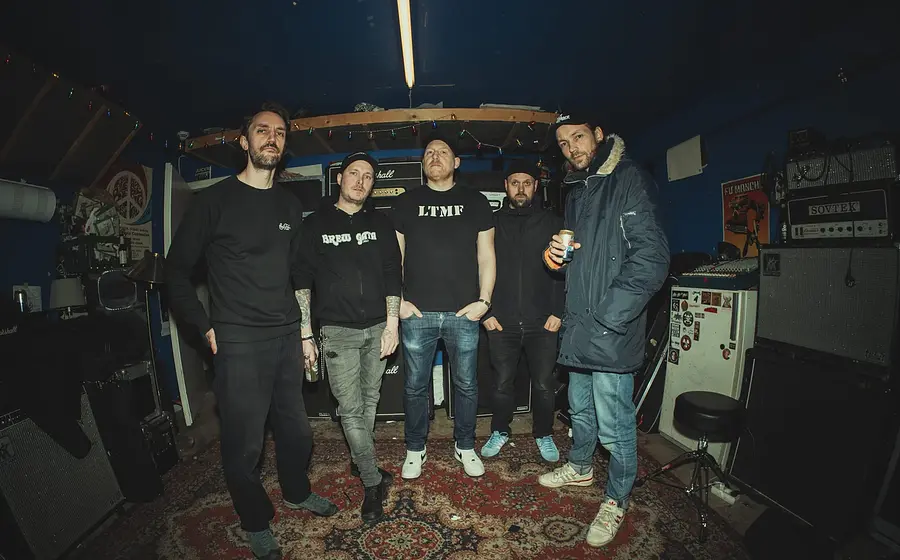 Bandfoto von The Good The Bad and The Zugly mit fünf Bandmitgliedern in dunkler Freizeitkleidung in einem Proberaum, umgeben von Verstärkern, Lautsprecherboxen und Musikequipment vor dunkelblauen Wänden.