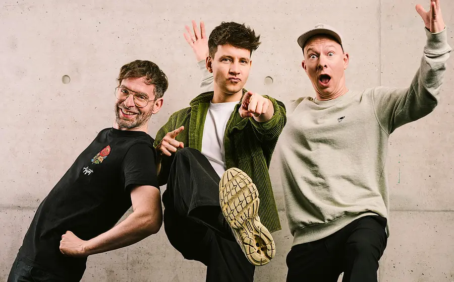 Die drei Bandmitglieder von Deine Freunde in Freizeitkleidung posieren vor einer hellen Mauer
