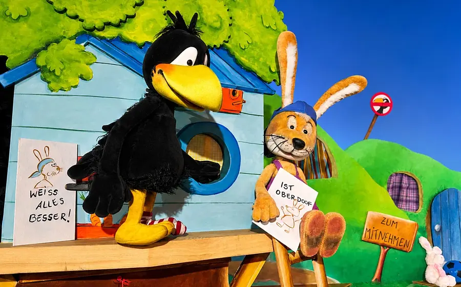 Bühnenfoto aus Der kleine Rabe Socke mit einer großen Rabenfigur und einer Hasenfigur vor bunten Häuschen und grüner Hügellandschaft, beide halten Schilder mit der Aufschrift Weiß alles besser! beziehungsweise Ist oberdoof!, daneben ein Schild mit der Aufschrift Zum Mitnehmen.