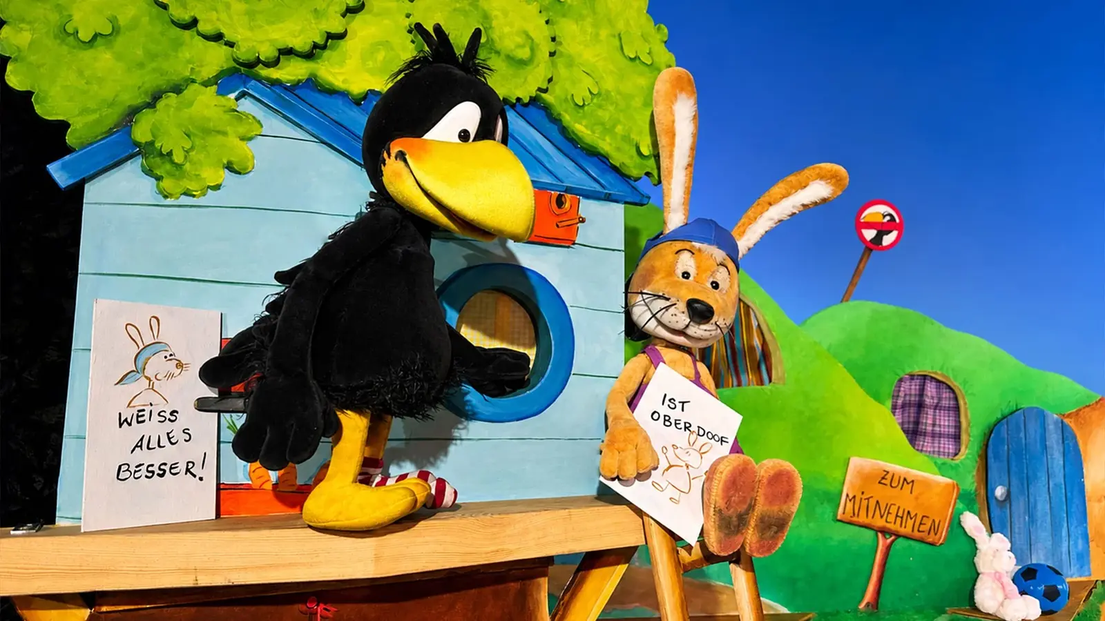 Bühnenfoto aus Der kleine Rabe Socke mit einer großen Rabenfigur und einer Hasenfigur vor bunten Häuschen und grüner Hügellandschaft, beide halten Schilder mit der Aufschrift Weiß alles besser! beziehungsweise Ist oberdoof!, daneben ein Schild mit der Aufschrift Zum Mitnehmen.