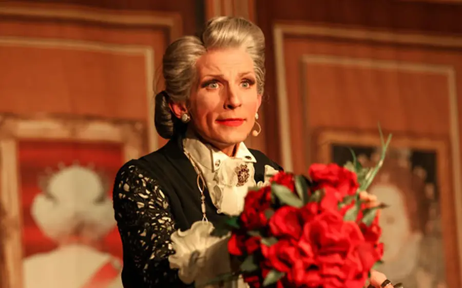 Person in historischer Bühnenkleidung und Hochsteckfrisur mit Blumenstrauß während einer Theateraufführung.