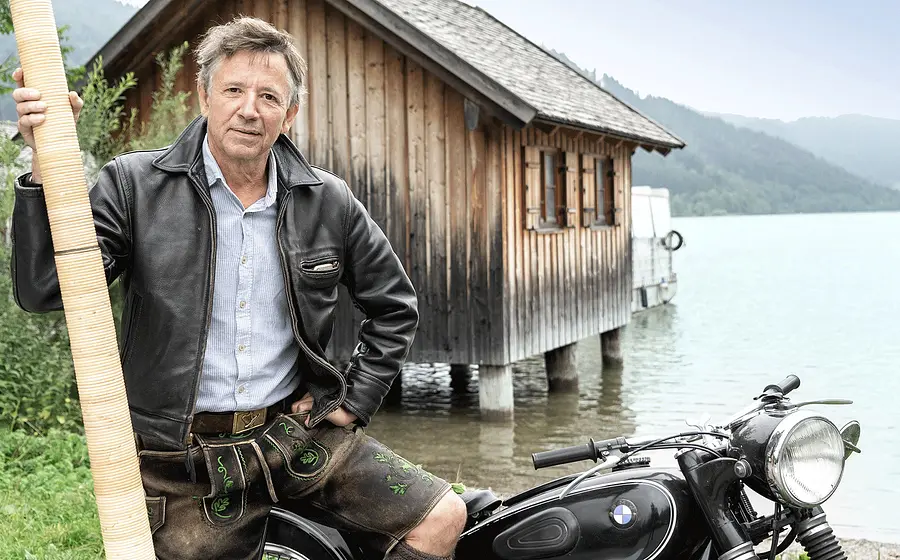 Person in Lederhose und Lederjacke neben einem historischen Motorrad am Ufer eines Sees und hält ein langes Alphorn. Im Hintergrund stehen ein Holzhaus auf Pfählen sowie bewaldete Berge am Seeufer.