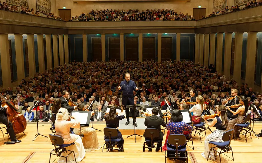 Orchester mit Dirigent vor großem Publikum in einem Konzertsaal