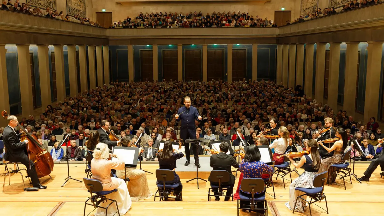Orchester mit Dirigent vor großem Publikum in einem Konzertsaal