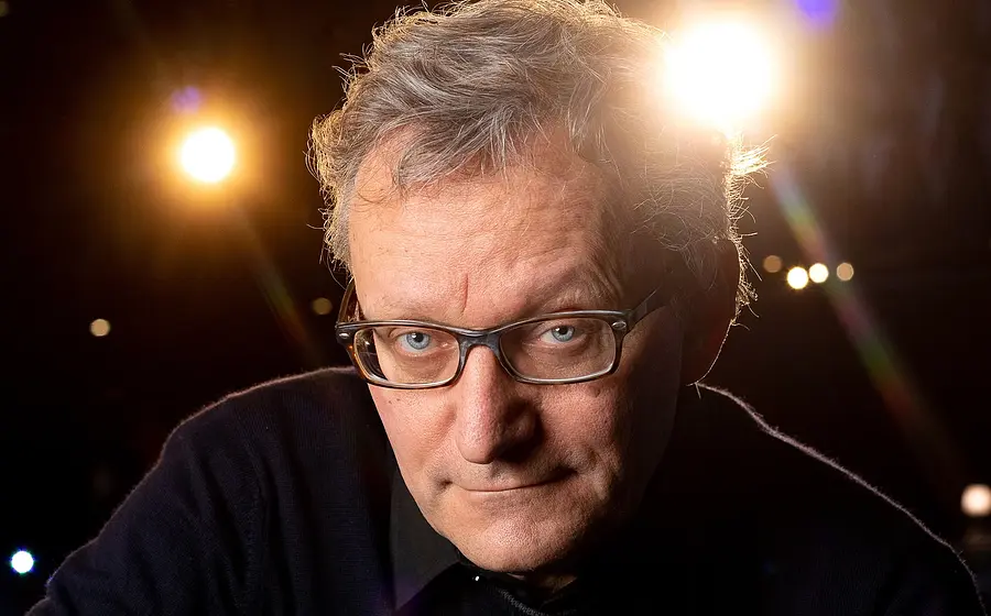Porträtfoto von Frank Lüdecke in dunklem Pullover und Brille vor unscharfem Bühnenhintergrund, mit direktem Blick zur Kamera und hellen Scheinwerfern im Hintergrund