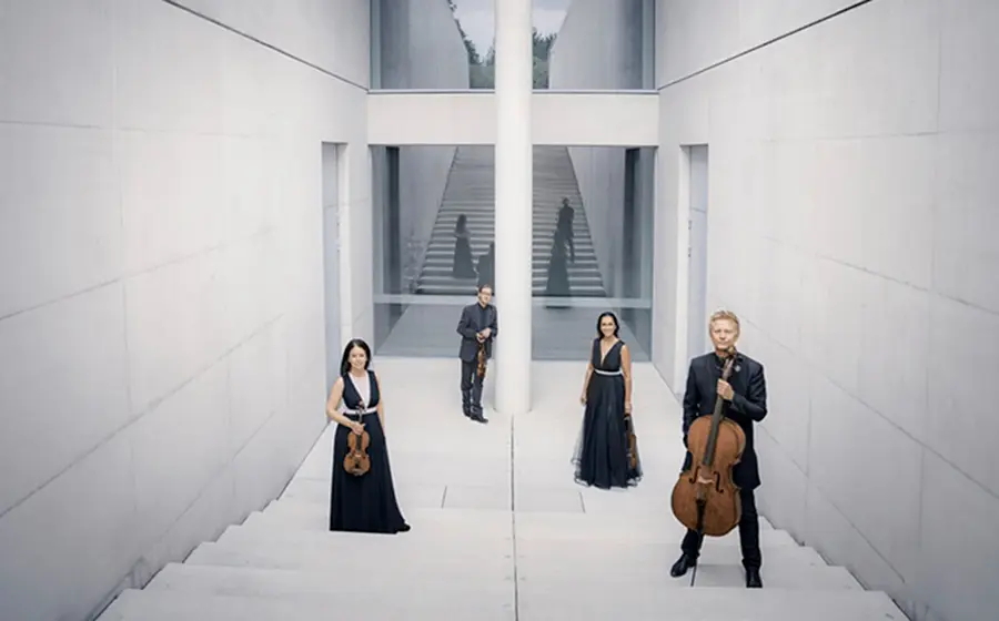 Streichquartett in einem modernen, hellen Treppenhaus. Die vier Ensemblemitglieder stehen mit ihren Streichinstrumenten auf einer breiten Treppenanlage vor klarer Architektur mit Treppen, Glasflächen und zentraler Säule