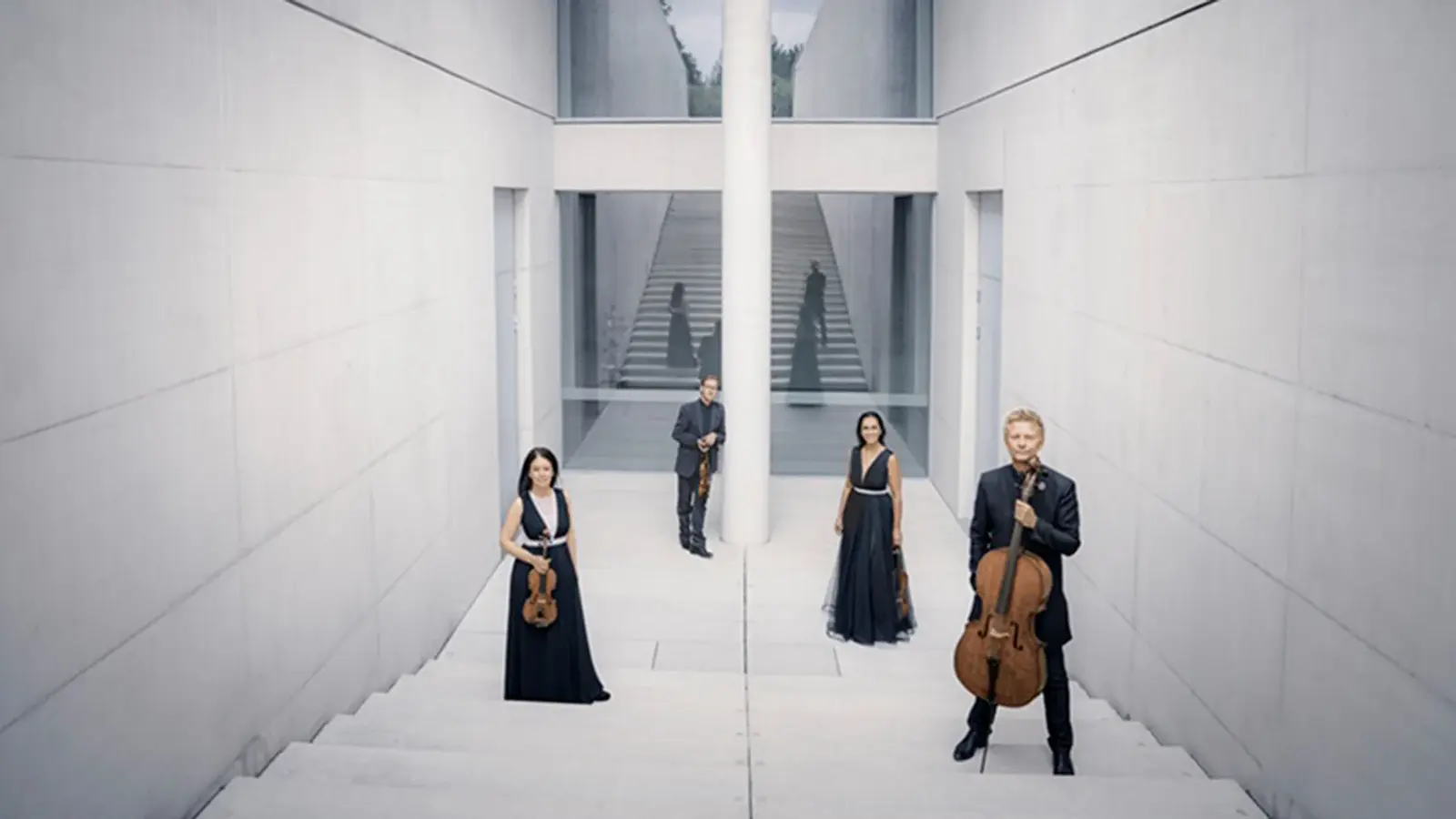 Streichquartett in einem modernen, hellen Treppenhaus. Die vier Ensemblemitglieder stehen mit ihren Streichinstrumenten auf einer breiten Treppenanlage vor klarer Architektur mit Treppen, Glasflächen und zentraler Säule
