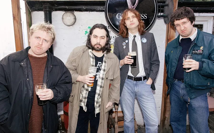 vier Personen stehen in legerer Kleidung vor einer weißen Außenwand mit rundem Guinness‑Schild, jede Person hält ein Getränk in der Hand. Tageslicht, informelle Atmosphäre; Motiv als Promo‑ und Eventbild der Band THE TUBS.