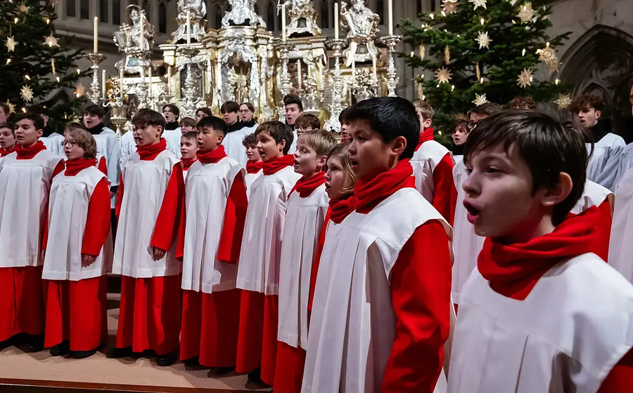 Bühnenfoto eines Knabenchors in roten und weißen Chorgewändern vor festlich geschmücktem Kirchenaltar mit Weihnachtsbäumen, Kerzen und goldfarbenem Altarschmuck im Hintergrund