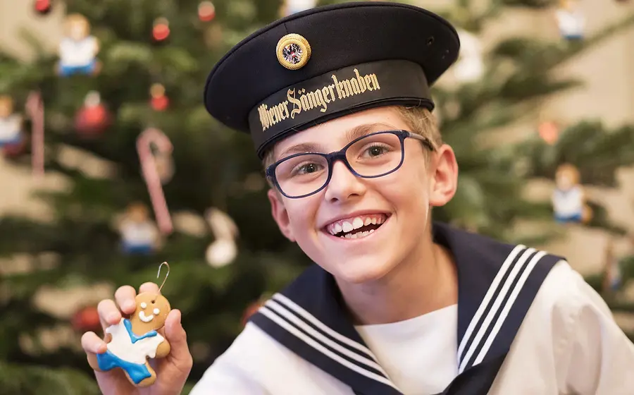 Porträtfoto eines Chorknaben der Wiener Sängerknaben in Matrosenuniform und Schirmmütze, mit Brille und Lebkuchenfigur in der Hand, vor festlich geschmücktem Weihnachtsbaum