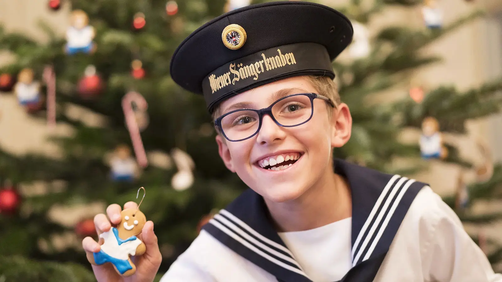 Porträtfoto eines Chorknaben der Wiener Sängerknaben in Matrosenuniform und Schirmmütze, mit Brille und Lebkuchenfigur in der Hand, vor festlich geschmücktem Weihnachtsbaum