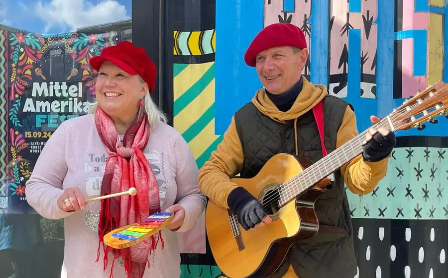 Foto mit zwei Personen vor bunter Wand mit grafischen Mustern und Plakat Mittel Amerika Fest eine Person mit Xylofon die andere mit Gitarre beide mit roten Mützen
