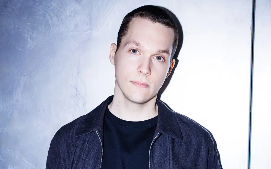 Ein Portrait von Lukas Sternath, der nachdenklich in die Kamera blickt, vor graublauem Hintergrund