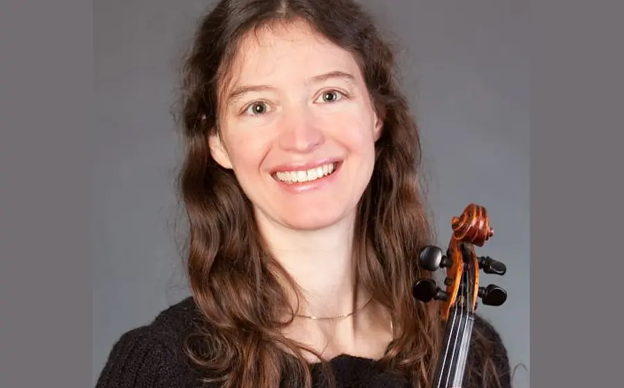 Porträtfoto einer Violinistin in schwarzem Oberteil vor grauem Hintergrund, mit langem brauen Haar und Violine an der Schulter, frontal zur Kamera blickend.