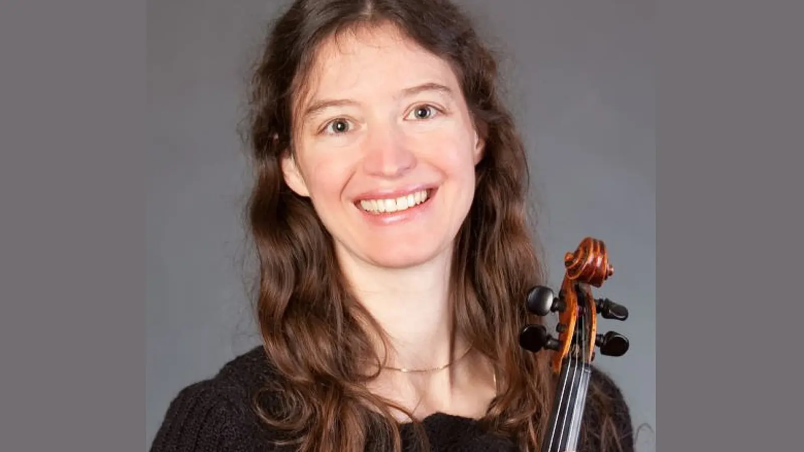Porträtfoto einer Violinistin in schwarzem Oberteil vor grauem Hintergrund, mit langem brauen Haar und Violine an der Schulter, frontal zur Kamera blickend.