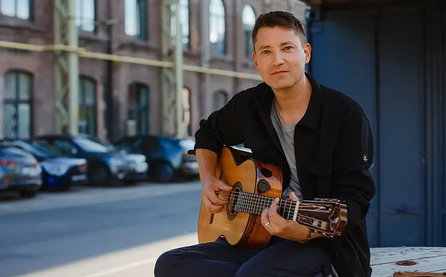 Daniel Stelter sitzend mit Gitarre in schwarzem Hemd, im Hintergrund eine Straßenszenen mit Hausfassade und parkenden Autos
