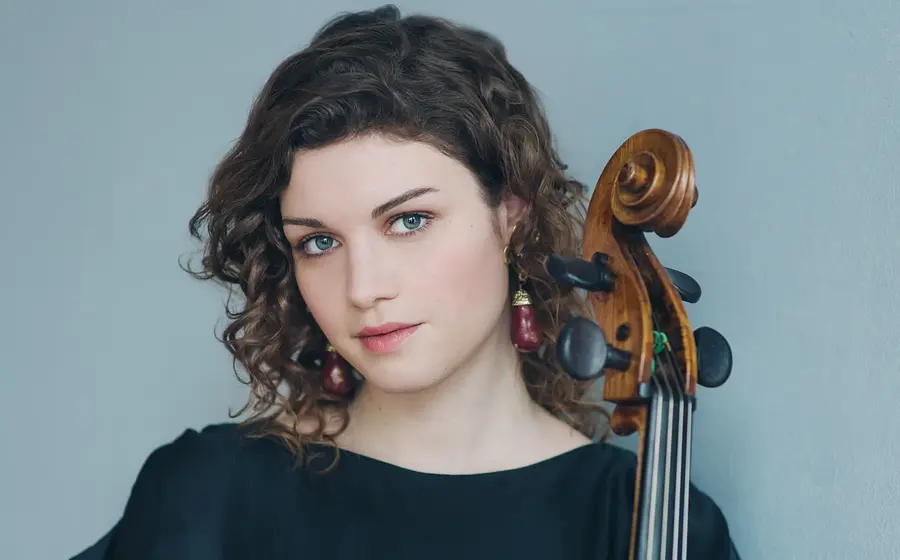Porträtfoto von Julia Hagen in schwarzer Kleidung, mit Violoncello neben dem Gesicht, vor hellblauem Hintergrund, mit lockigem Haar und rotem Ohrschmuck.