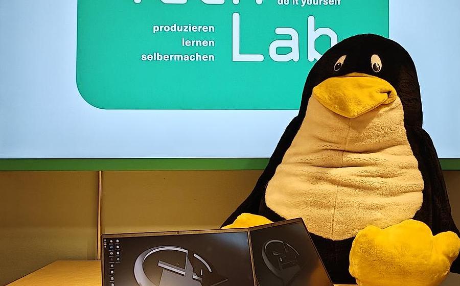 Ein Stoff-Pinguin sitzt auf einem Tisch, davor Elektroschrott.