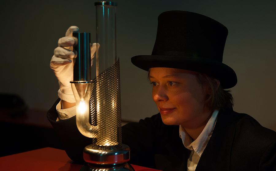 Eine Frau in schwarzem Anzug, mit weißen Handschuhen und mit schwarzem Zylinder experimentiert in einem abgedunkelten Raum. Auf der roten Tischplatte vor ihr steht ein Glasrohr, aus dem ein weiteres Glasrohr abzweigt wie ein Arm eines Kaktus. Auf diesem Rohr sitzt ein silbernes metallisches Rohr, darunter brennt eine kleine Flamme, etwa so groß wie von einer Kerze. Die Frau hält das silberne Rohr in einer Hand und schaut gespannt auf die Flamme.