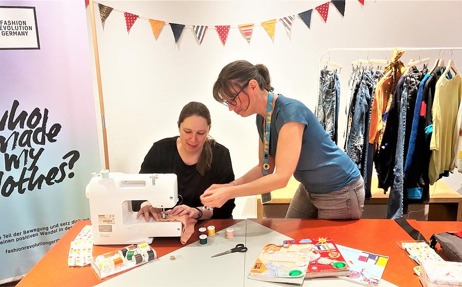 Zwei Frauen reparieren Textilien mit einer Nähmaschine. Im Hintergrund ein Kleiderständer mit verschiedenen Textilien.