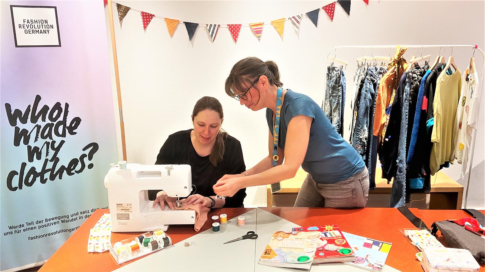 Zwei Frauen reparieren Textilien mit einer Nähmaschine. Im Hintergrund ein Kleiderständer mit verschiedenen Textilien.