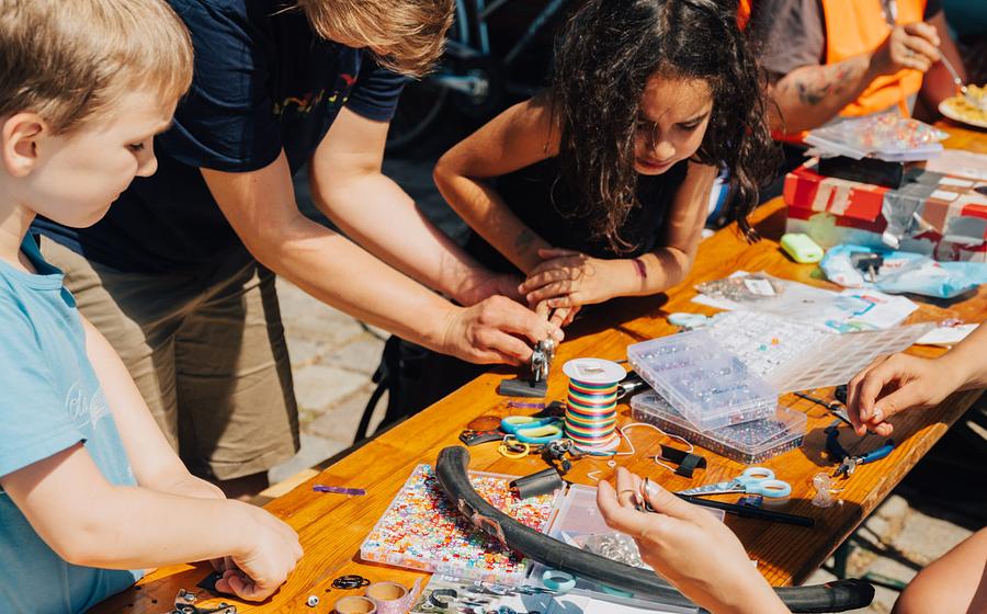 Mehrere Erwachsene und Kinder sitzen und stehen um einen Biertisch im Freien. Auf dem Tisch liegen verschiedene Bastelmaterialien. Die Personen basteln gemeinsam. Im Hintergrund steht ein Fahrrad.
