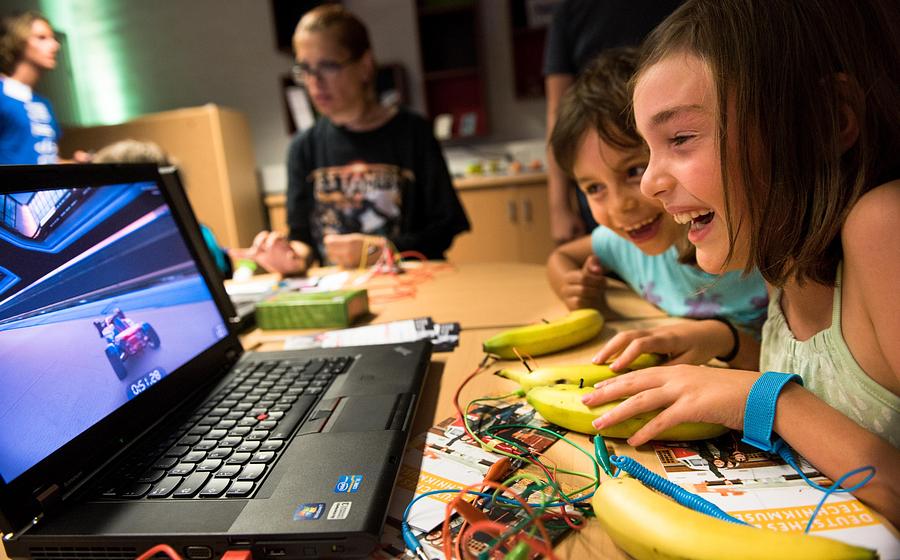 Kinder sitzen an einem Tisch vor einem Laptop. Auf dem Bildschirm ist ein Spiel mit einem Rennauto zu sehen. Die Kinder steuern das Spiel mit Bananen, die auf dem Tisch liegen und über Kabel und ein Gerät mit dem Laptop verbunden sind.