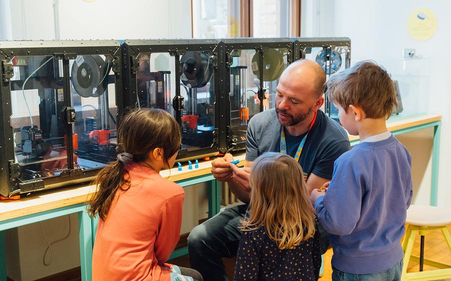 Ein Mann und drei Kinder sitzen vor einer Reihe von 3D-Druckern. Der Mann erklärt etwas, die Kinder haben Spaß und hören zu.
